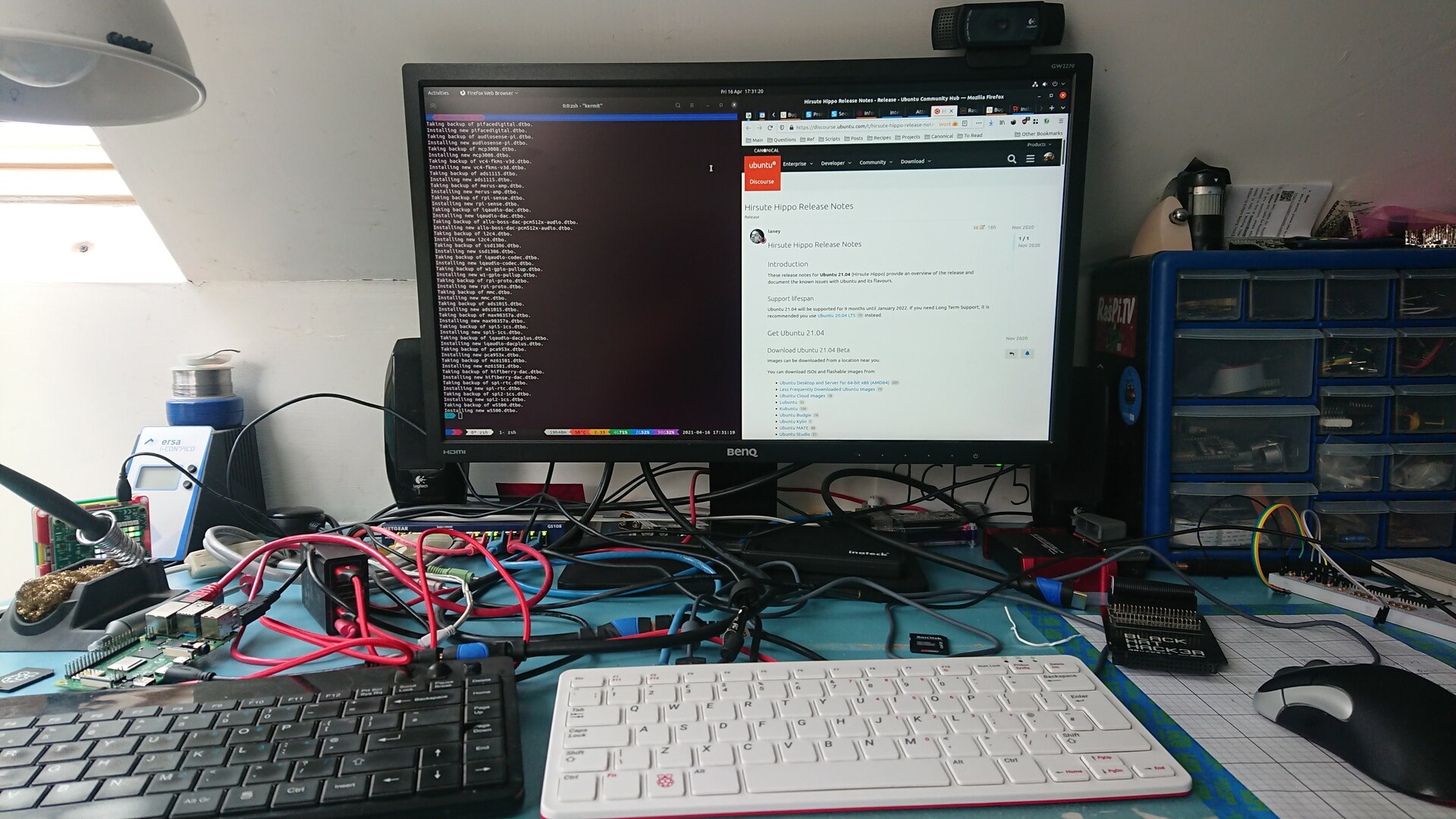 A Pi 400 in front of a simple HDMI monitor with a Logitech C920 webcam attached, and a USB3 SSD resting on the base, surrounded by the typical junk on Dave's desk (myriad cables, a solder station, a spare keyboard for testing, random SD cards)