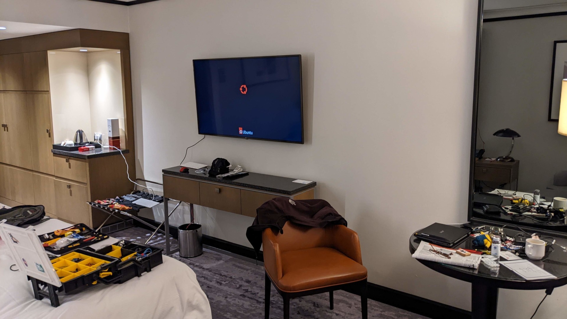 An image of Dave's hotel room at an engineering sprint. Amidst a collection of socks, a Pi 400 is hooked up to the huge 40-something inch TV in front of the bed. The screen is displaying the Ubuntu logo typically seen during desktop boot. In the left foreground, two toolboxes are open on the bed. On the right, a small table is covered in the detritus of the day: Dave's laptop, empty coffee mug, glasses, a copy of Private Eye, a graphic novel, and several random bits of Pi paraphenalia.