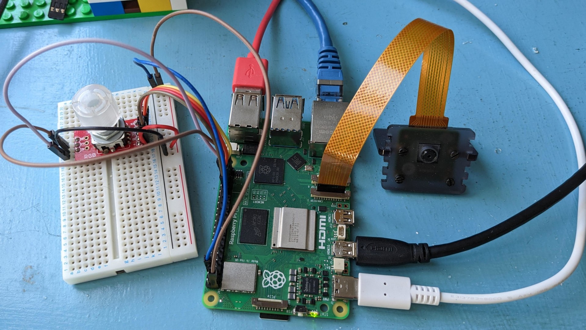 A Raspberry Pi 5 sits on top of a blue-painted desk. Various cables (USB, ethernet, micro-HDMI, and USB-C power cable) are plugged into it, as well as a brown ribbon cable (FFC) leading to a Raspberry Pi camera module. To the left, various jumper leads trail from the GPIO header to a small breadboard containing a rotary encoder with an RGBLED embedded within it.